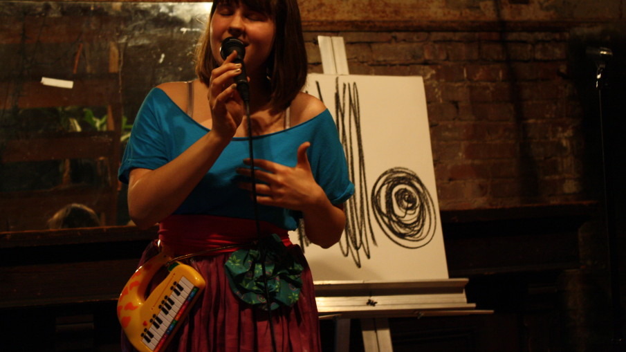 A young Alexa Dexa with a short bob haircut sings into a microphone with her live-drawn charcoal art behind her on an easel and a belt holding her electronic giraffe toy piano.