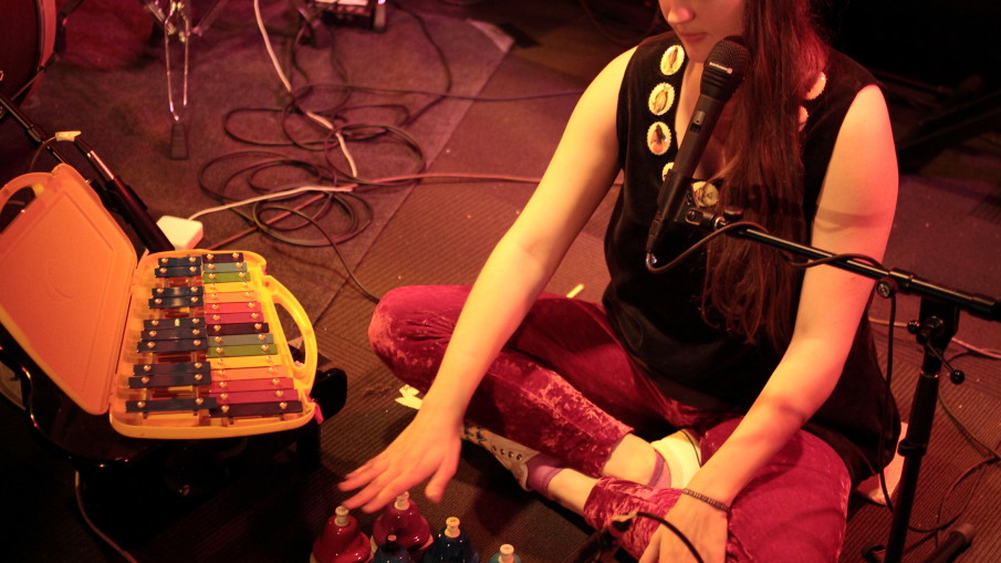 Alexa Dexa sits on the floor with their toy piano, toy glockenspiel, and pitched desk bells to play a live shows in their red velvet pants and velvet dress with a collar of buttons with fishes on them.