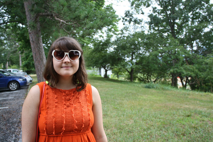 Alexa Dexa in an orange dress with heart-shaped sunglasses at a sculpture park.