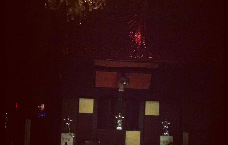 Alexa Dexa plays a live show with their toy piano and desk bells in a dim room with a chandelier.