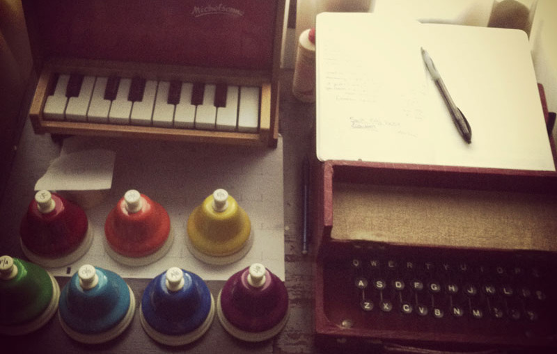 A collection of toy instruments including a trombone kazoo, toy piano, desk bells, and typatune as well as a notebook and a pen.