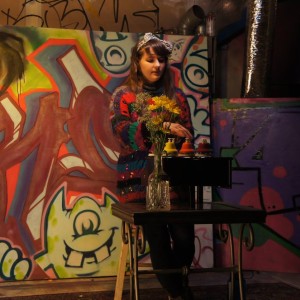 Alexa Dexa plays a live show with their toy piano and pitched desk bells in front of a wall of graffiti art.