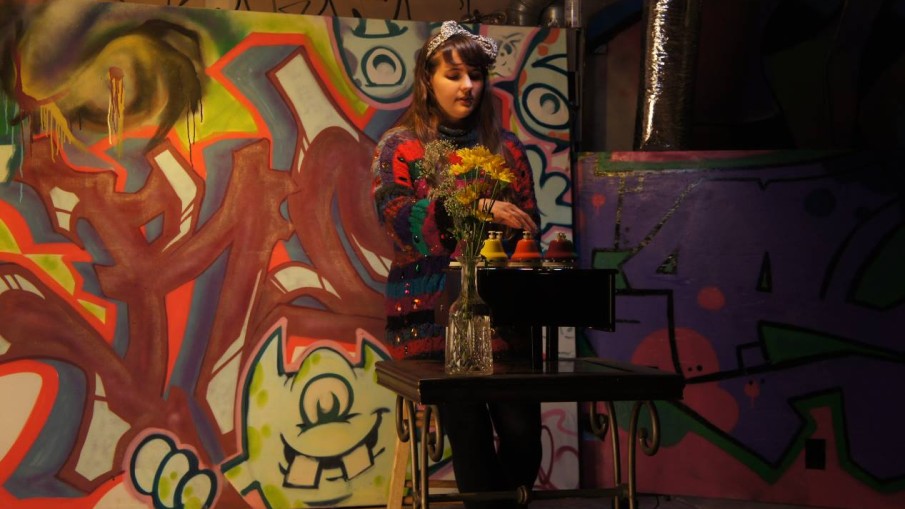 Alexa Dexa plays a live show with their toy piano and pitched desk bells in front of a wall of graffiti art.