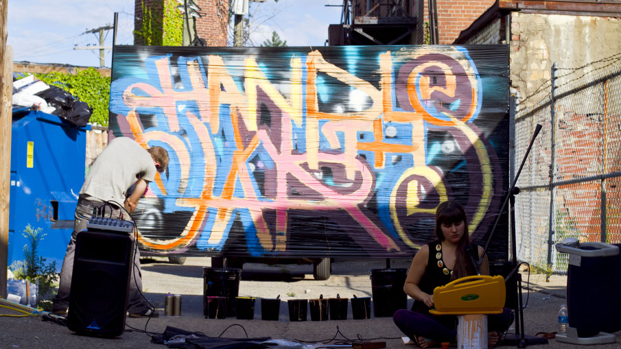 Alexa Dexa plays her toy glockenspiel and found objects at Detroit's Sidewalk Festival of Performing Arts. Artist Kevin McCoy live-paints behind them.