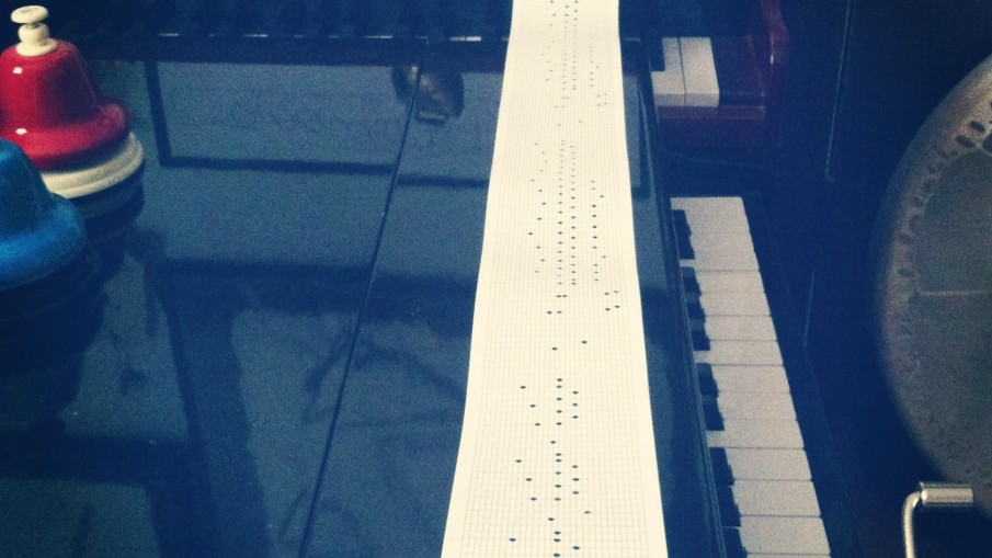 Sitting on top of a baby grand toy piano is a hand-cranked music box with a hand-punched music scroll being cranked through.