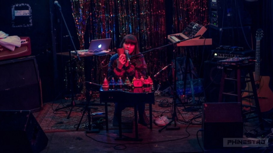 Alexa Dexa sits at her baby grand toy piano holding her kalimba in her hands with their pitched desk bells sitting on top of their toy piano at a live performance with silver streamers in the background.