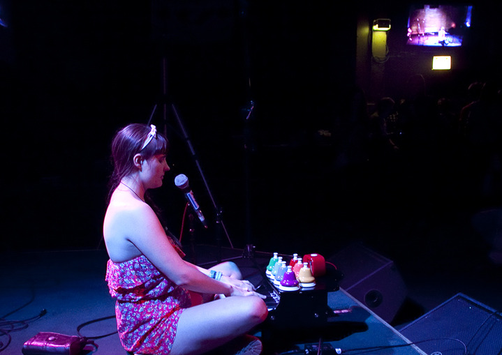 A profile view of Alexa Dexa sitting on the floor playing their toy piano and pitched desk bells on stage at a live show.
