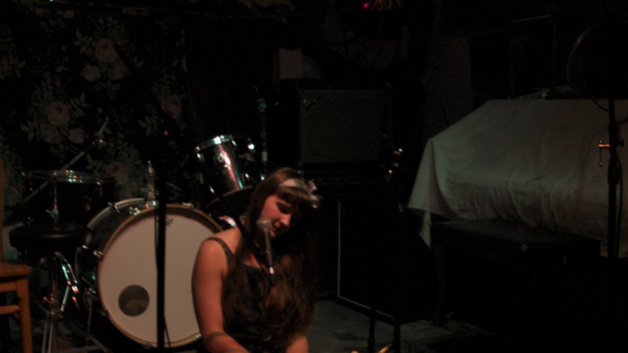 In a dimly lit venue, Alexa Dexa sits on the floor playing their toy piano and desk bells at a performance.