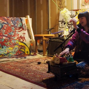 Alexa Dexa sits on the floor with their hands floating above their desk bells, hitting them and keeping time in fluid motions at a live show among paintings.