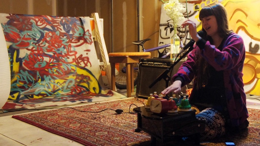 Alexa Dexa sits on the floor with their hands floating above their desk bells, hitting them and keeping time in fluid motions at a live show among paintings.