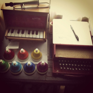 A collection of toy instruments including a trombone kazoo, toy piano, desk bells, and typatune as well as a notebook and a pen.