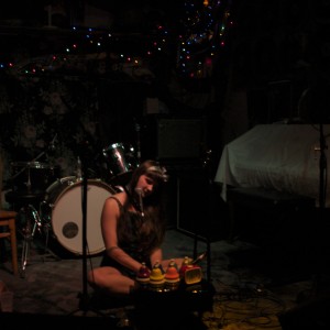 In a dimly lit venue, Alexa Dexa sits on the floor playing their toy piano and desk bells at a performance.