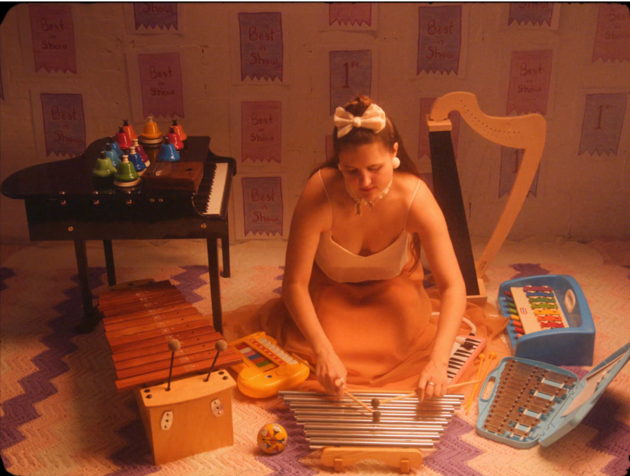 alexa dexa, in a silk gown with a white top and flowing peach skirtlength, kneels on the floor playing their toy chimes, Surrounding them from left to right are their 37-key Schoenhut toy piano, 20 chromatic desk bells, kalimba, alto xylophone, toy glockenspiel, tap-a-tune xylo-keyboard, and toy harp.