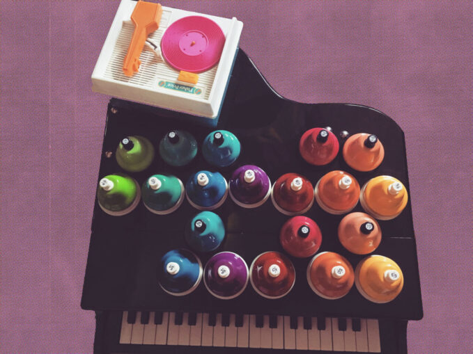 Alexa Dexa’s 37 key @schoenhutpiano baby grand toy piano floats on a purple background. On top of the toy piano sit a very colorful set of 20 chromatic toy desk bells and a toy record player with a custom pink 3D printed toy record which plays a tune Alexa composed for their electroacoustic toy opera, Be a Doll.