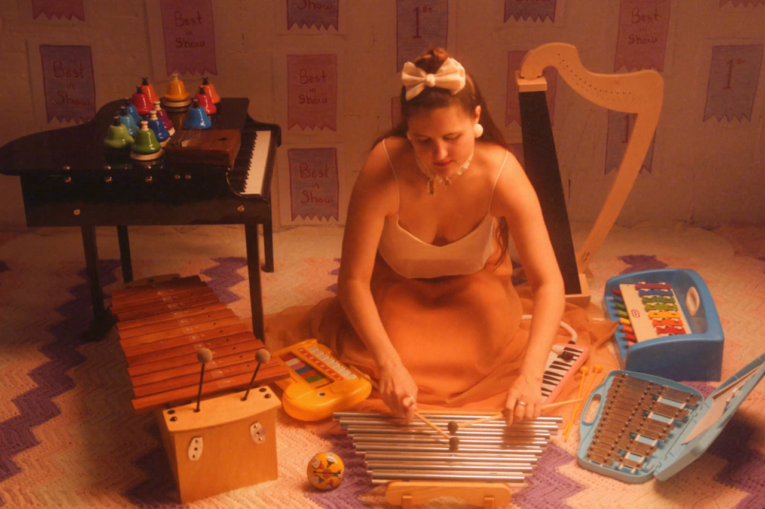 alexa dexa, in a silk gown with a white top and flowing peach skirtlength, kneels on the floor playing their toy chimes, Surrounding them from left to right are their 37-key Schoenhut toy piano, 20 chromatic desk bells, kalimba, alto xylophone, toy glockenspiel, tap-a-tune xylo-keyboard, and toy harp.